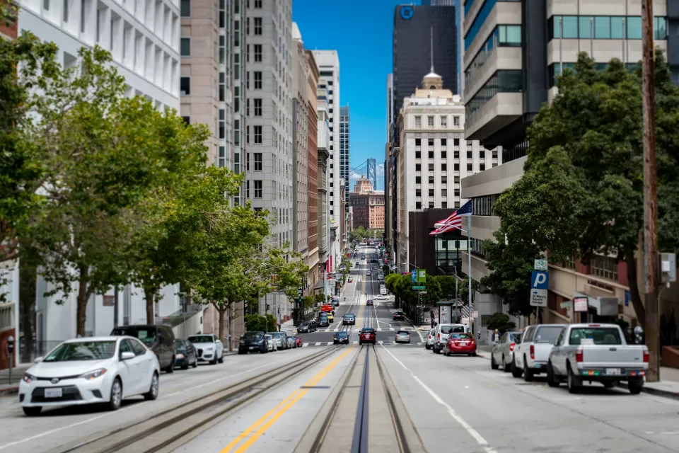 View from middle of street with streetcar rails and high-rise buildings on either side in San Francisco, Bay Bridge visible in background